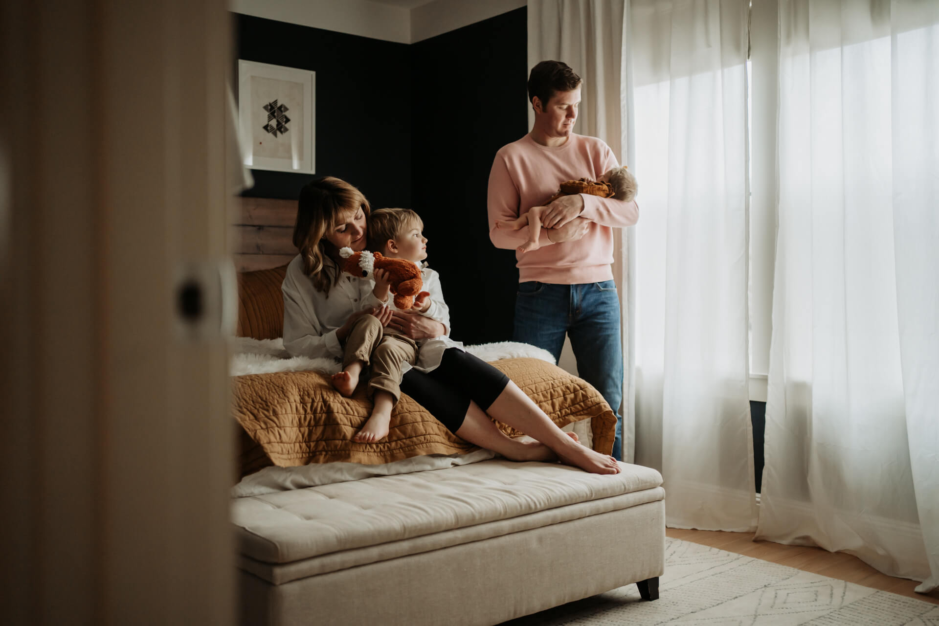 Mom and toddler sitting on a bed while dad is holding his newborn baby girls by the window