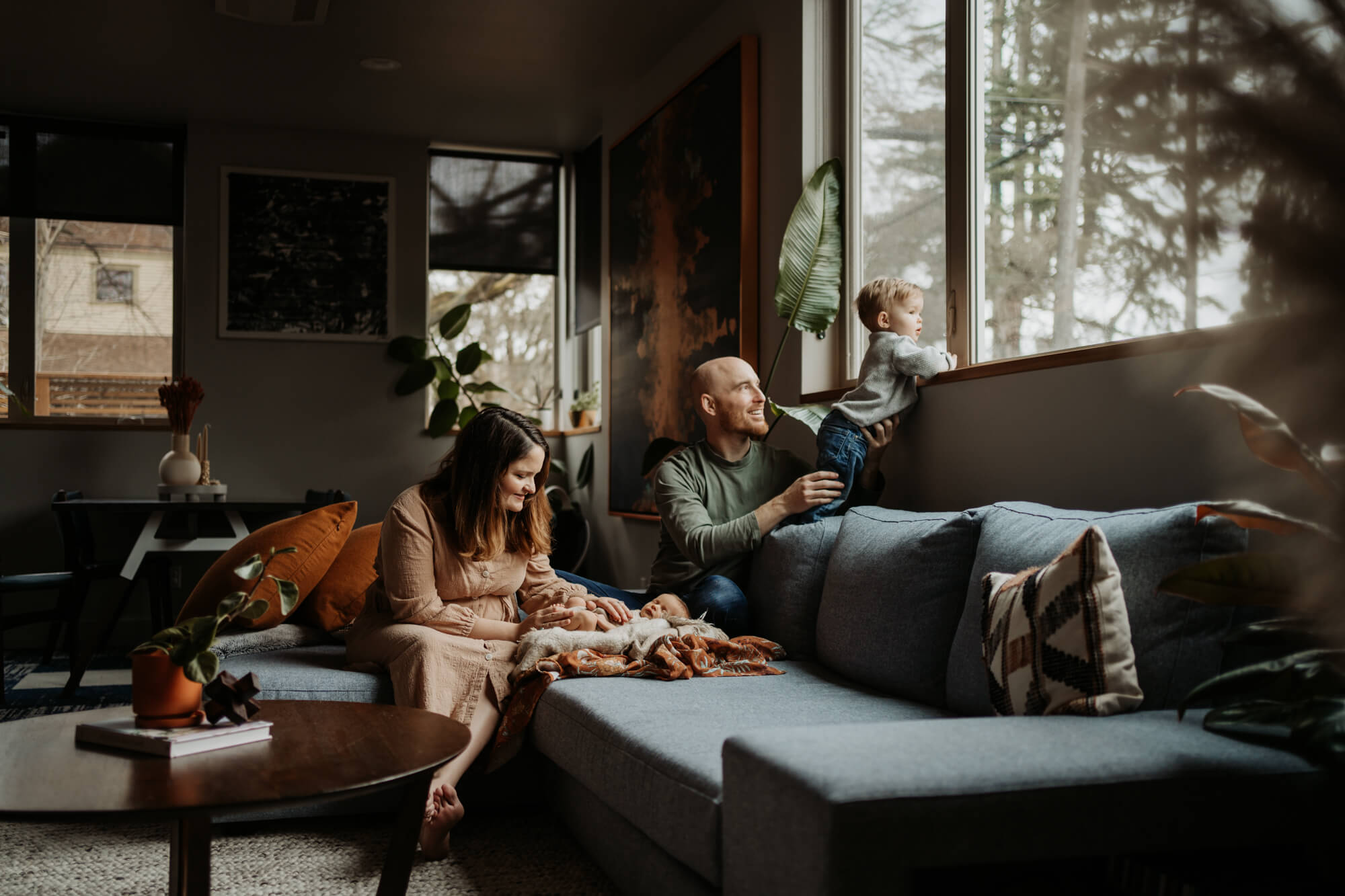 Family of four relaxing on their living room as mom looking at her newborn baby and dad and toddler boy looking out for the window