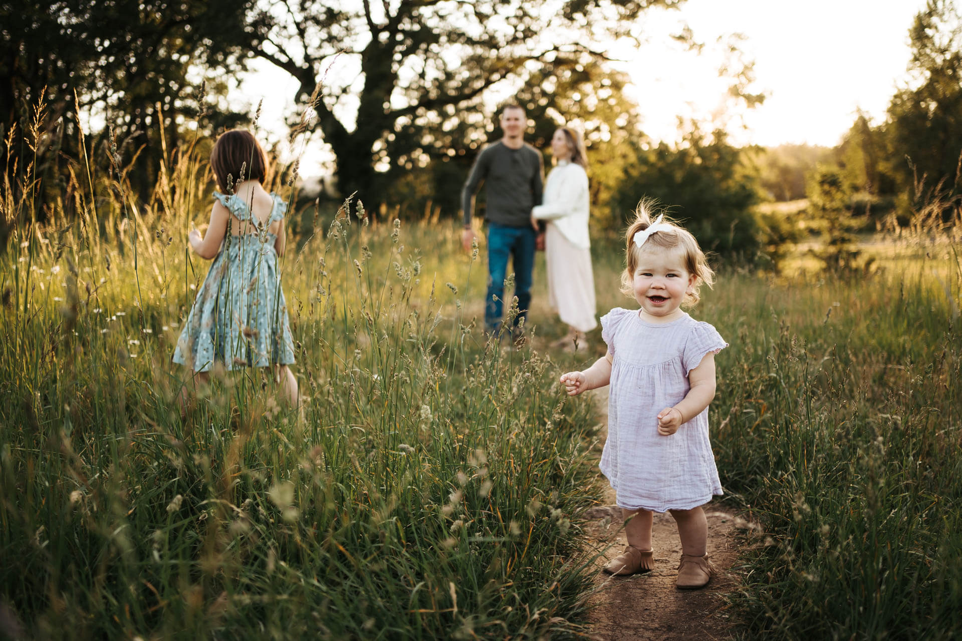 Toddler girl is laughing on a flower field while her big sister and parents stand in the same field'