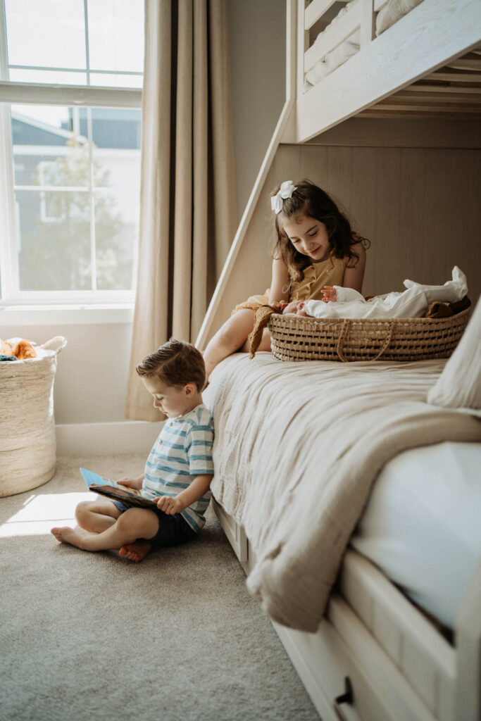A toddler girl touching baby sister's head on the bed while a toddler boy reading a book by the bed