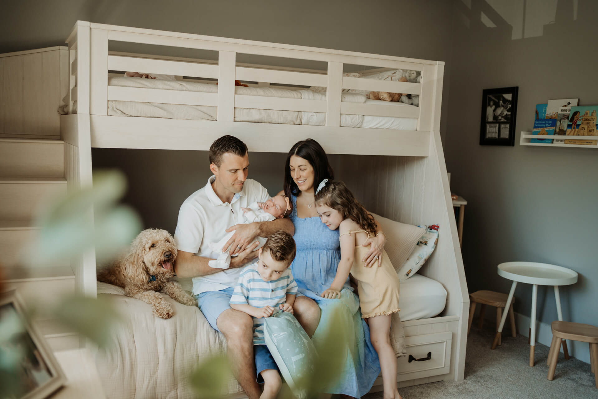 Dad holding his newborn baby girl while sitting on a bed and 2 toddlers are standing by their mom who is sitting right next to the dad
