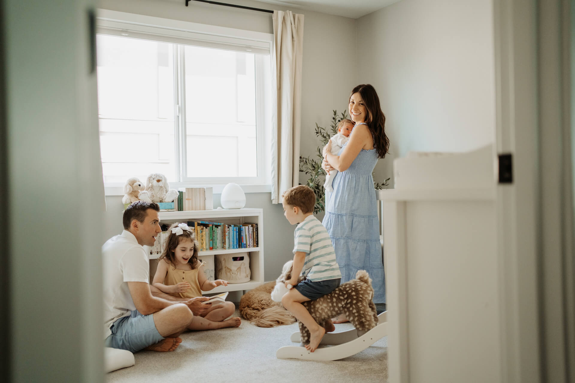 Dad and his toddler girl reading a book while a toddler boy riding his horse toy. Mom is holding newborn baby and smiling at a camera