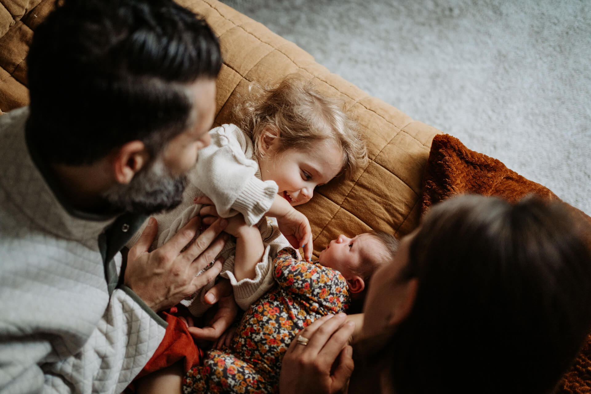 Toddler girl and newborn baby girl looking at each other as they both lay on a bed while the parents looking at their children