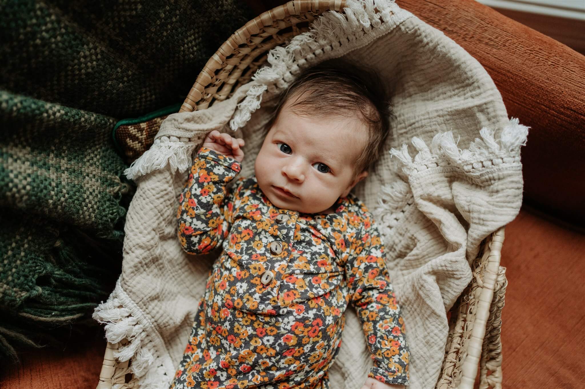 Newborn baby girl in floral onesie is laying in a wicker bassinet and looking at a camera