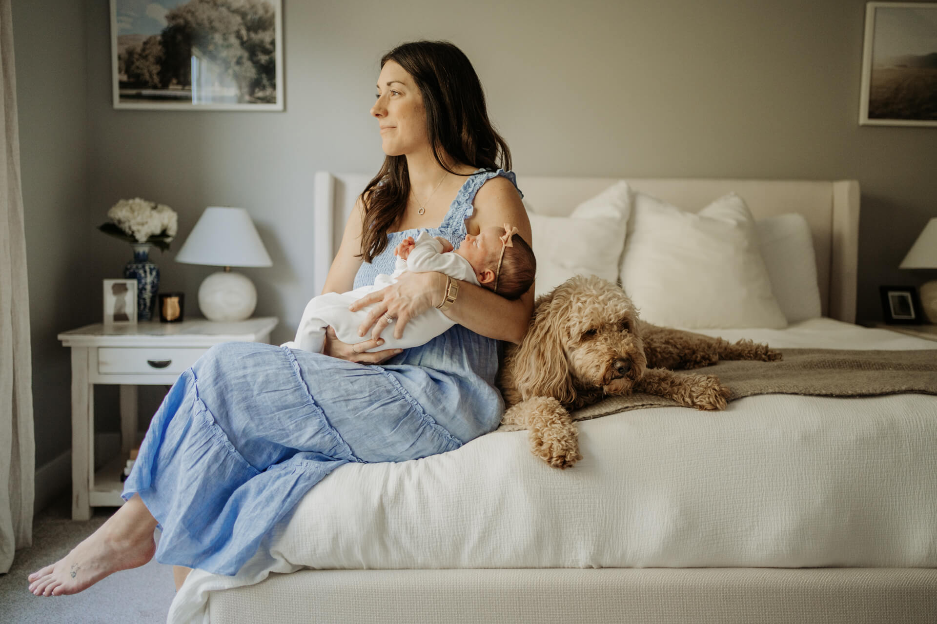 Mom holding her baby in her arm while sitting on her bed. A golden-doodle puppy is laying down right next to them on the bed. 