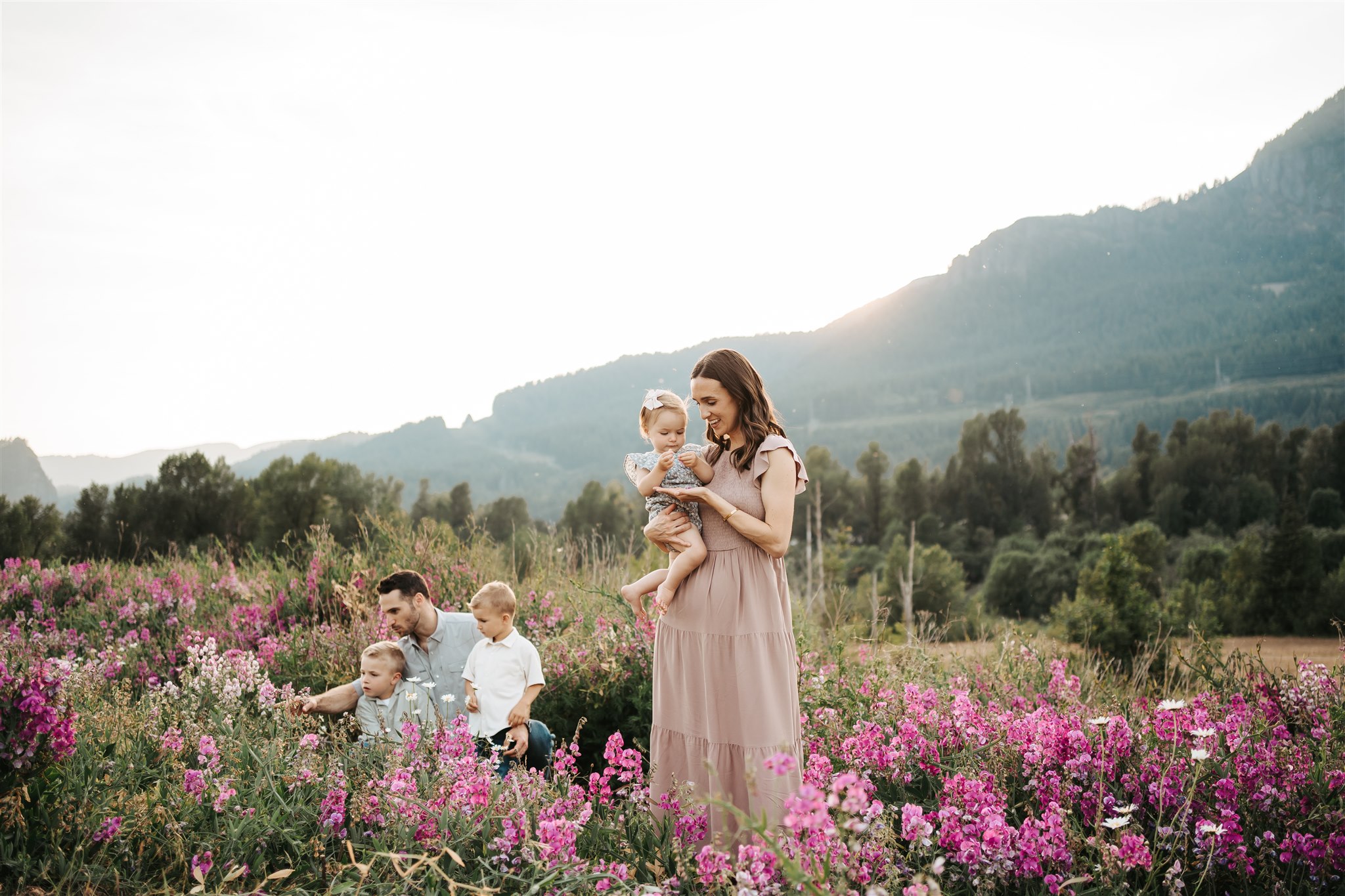Mom standing and holding a baby while dad is looking at pink flowers with 2 of his toddlers